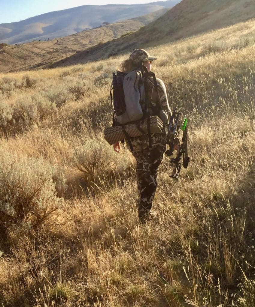 Photo of Jessi Johnson, a new hunter, walking through field with hunting pack and bow.